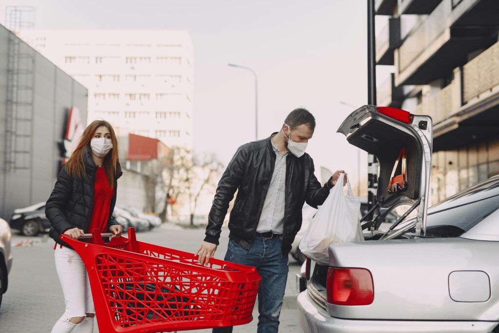 Couple in casual clothes and protective masks moving shopping bags from supermarket trolley to car trunk on street near modern multistory buildings